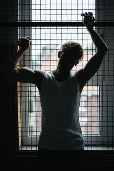 Silhouette of male athlete in casual clothes standing against light and holding on metal lattice