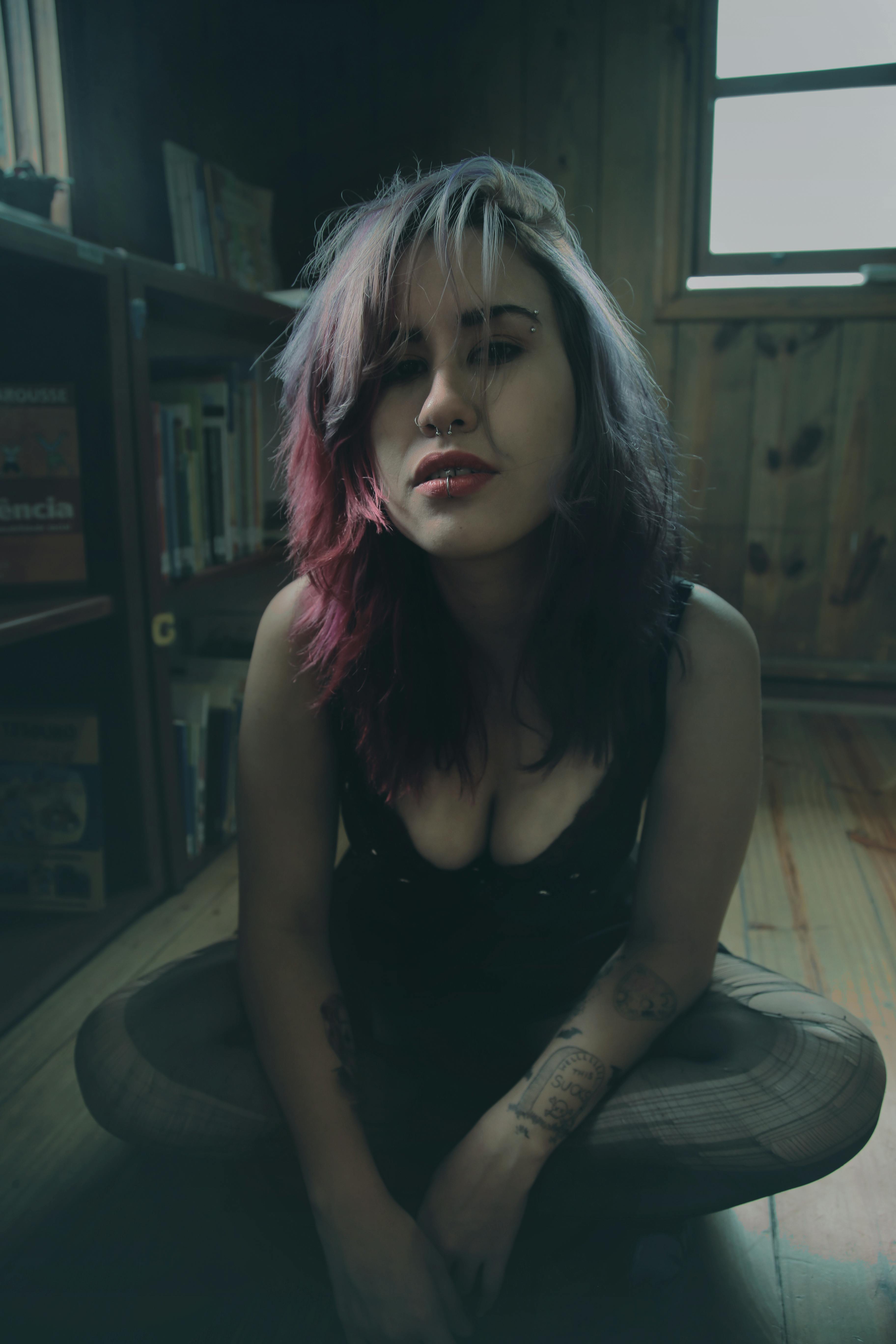 Moody portrait of a young woman with piercings and pink hair sitting in a wooden room.