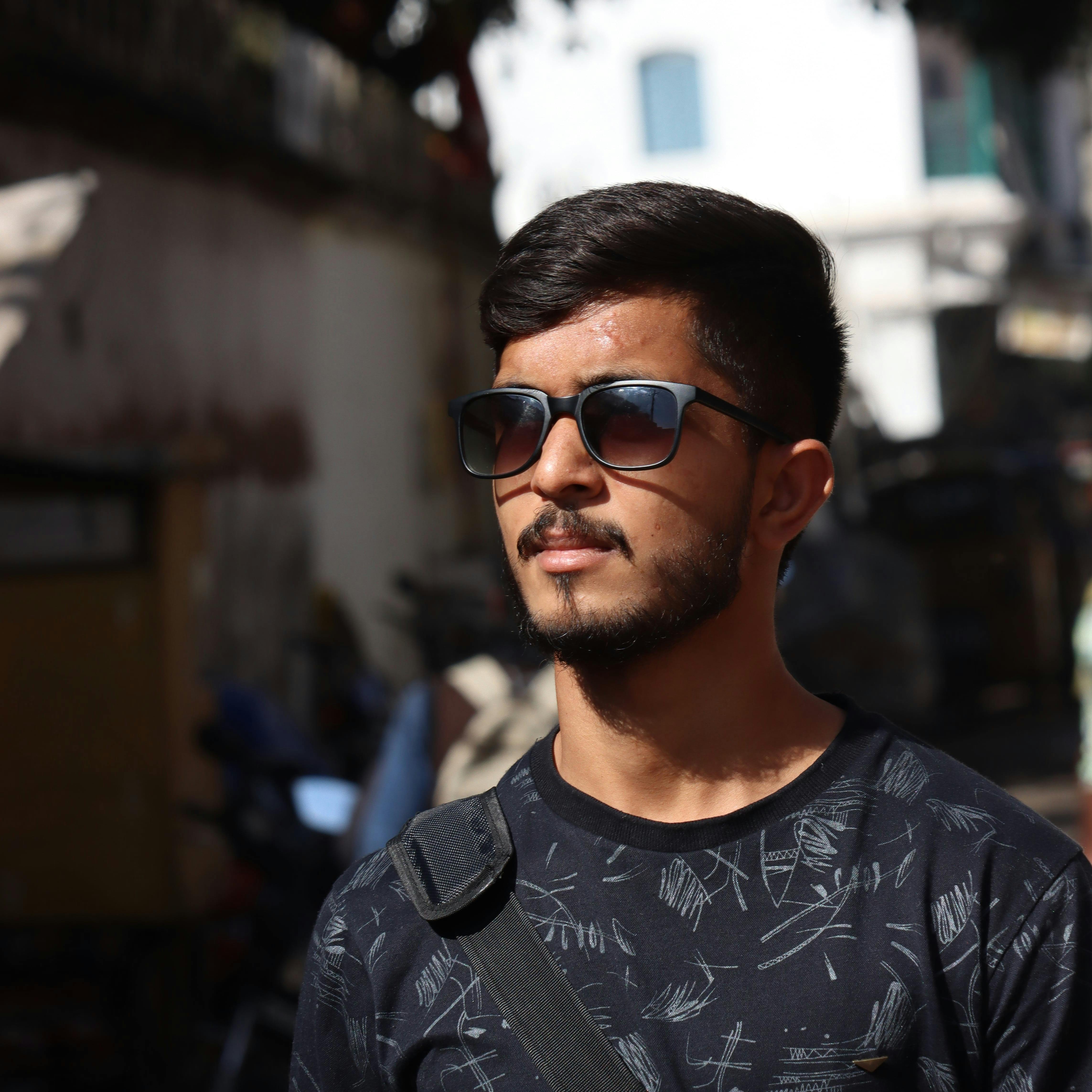 A fashionable young man with sunglasses stands confidently in the sun, showcasing urban style.