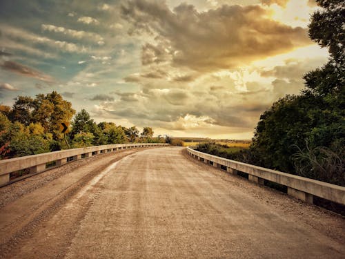 Free Gray Concrete Road Between Green Trees Under White Clouds and Blue Sky Stock Photo