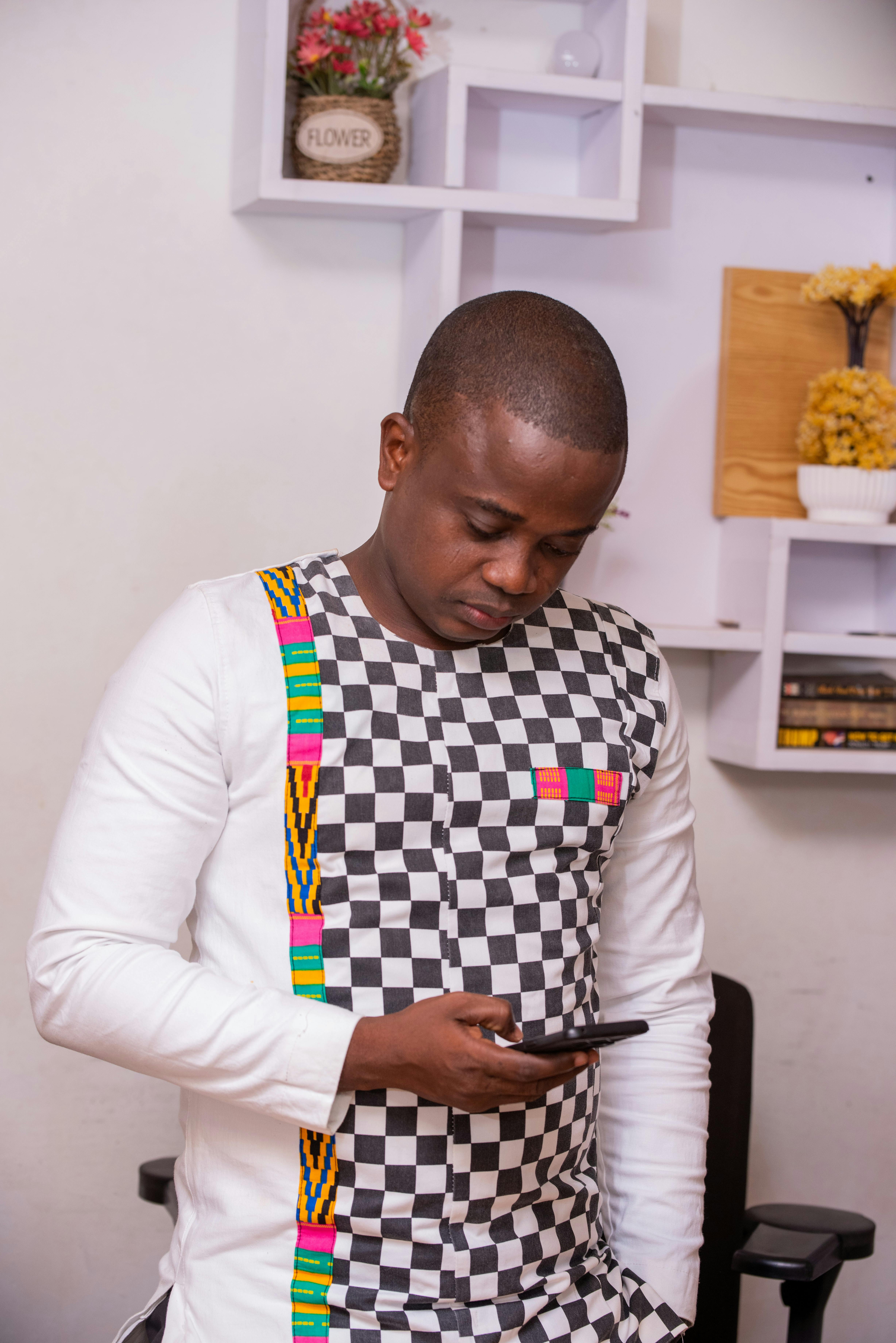 Young African man in traditional Ghanaian attire using a smartphone indoors.