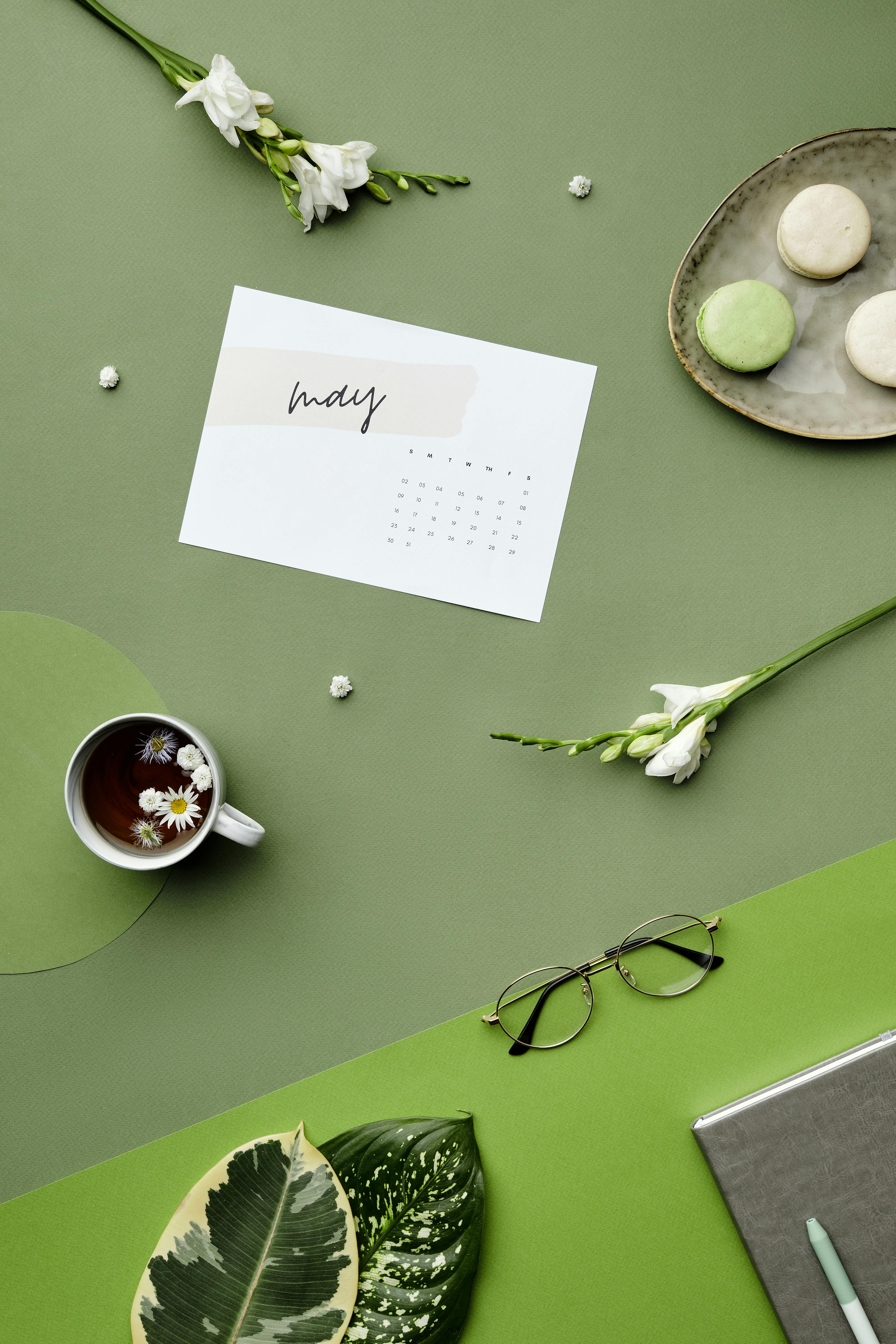 A chic flat lay featuring a May calendar, tea, flowers, and macarons on a green background.
