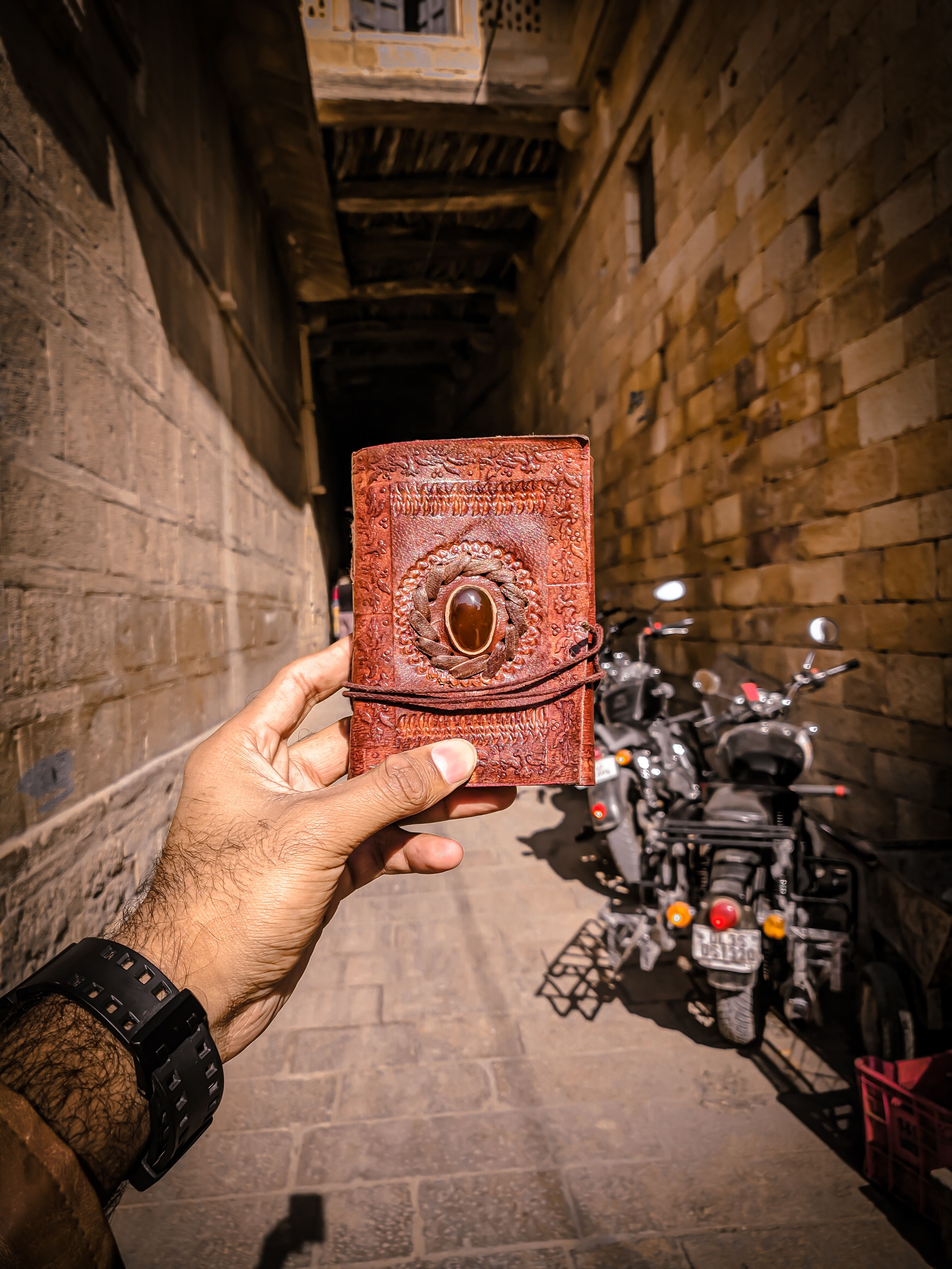 A hand holds a vintage leather journal on a sunny street lined with stone walls and motorcycles in Jaisalmer.