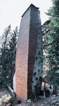 Aged masonry tower on dry land near overgrown green trees under white sky in town