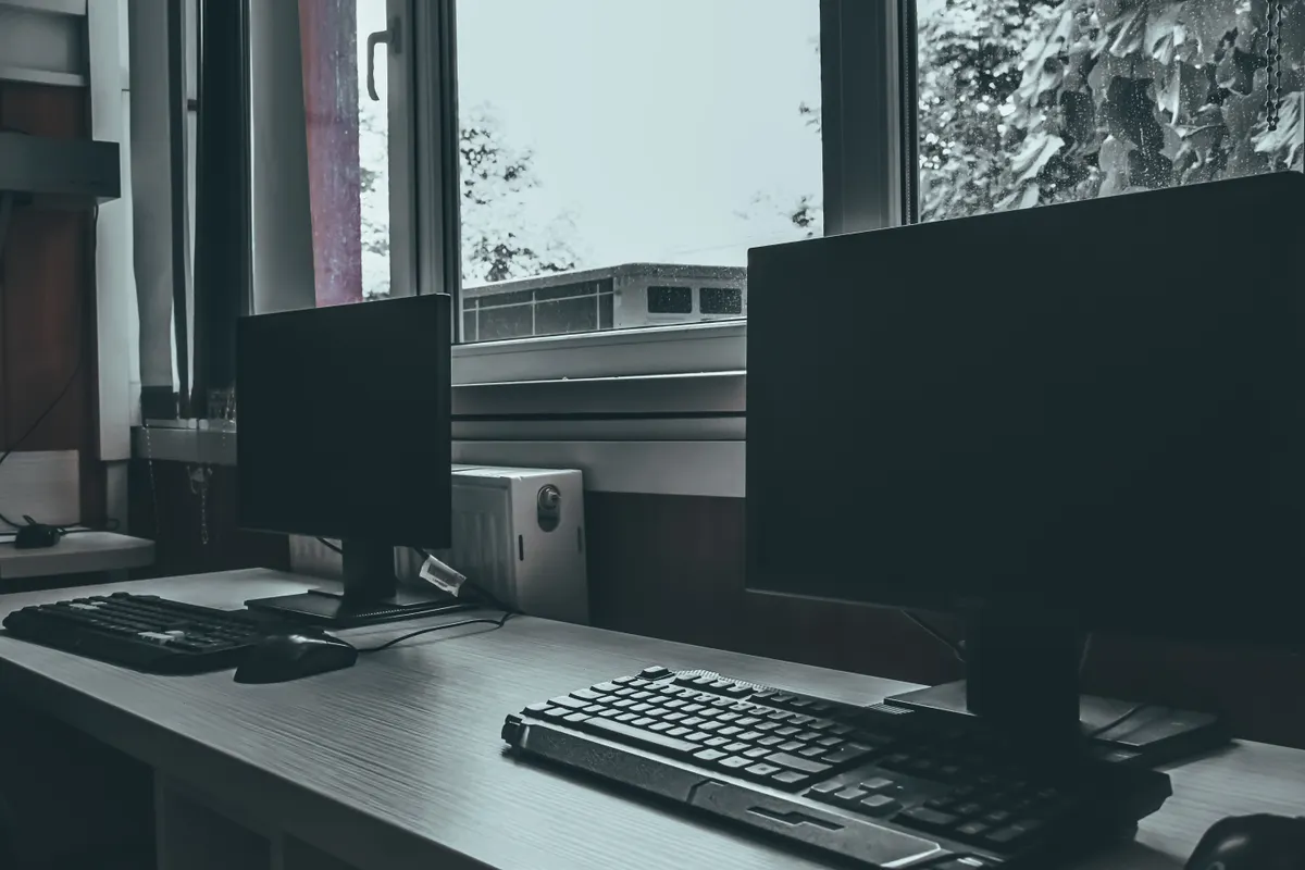 An empty desk beside a window with two dark, switched-off monitors and their keyboards.