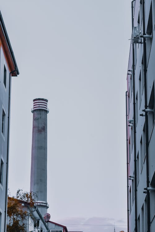 Low angle of tall smokestack pipe near simple buildings in industrial district against foggy sky