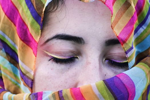 Calm young ethnic female with eyes closed and bright eyeliner on eyelids in vibrant multi colored headscarf in daylight