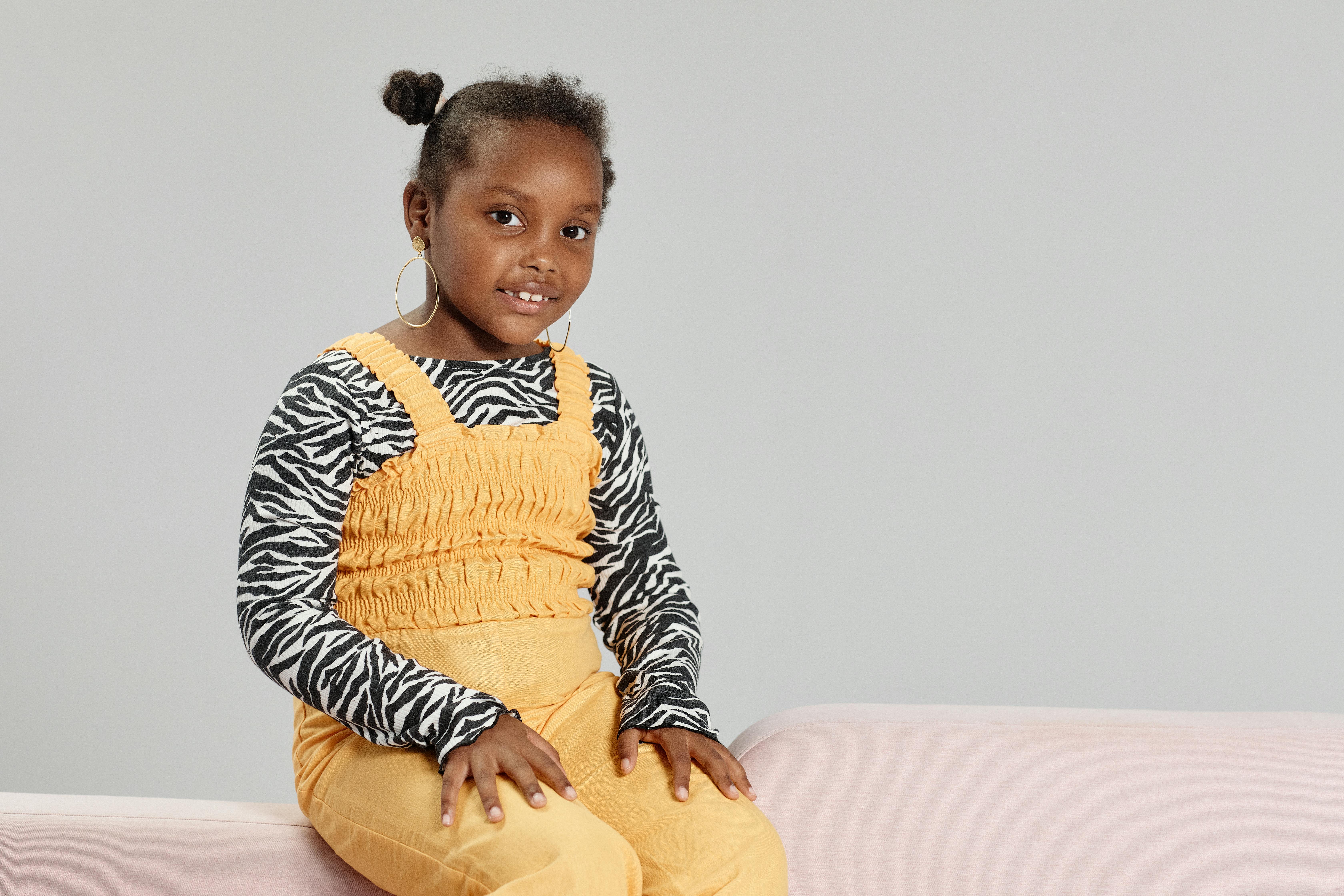 A young girl poses stylishly in a zebra print top and mustard romper, featuring hoop earrings.