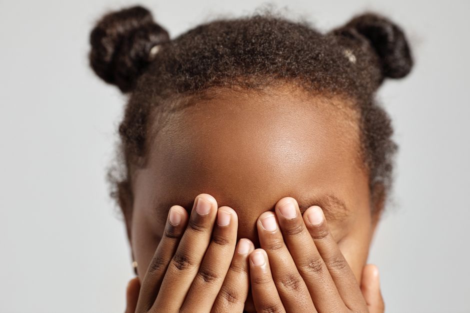 Close-up portrait of a child covering their eyes with hands, posing in a studio setting with a neutral background.