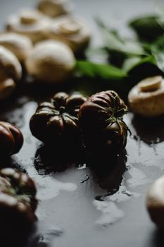 Wet mushrooms and heirloom tomatoes arranged on a dark surface with leafy greens.