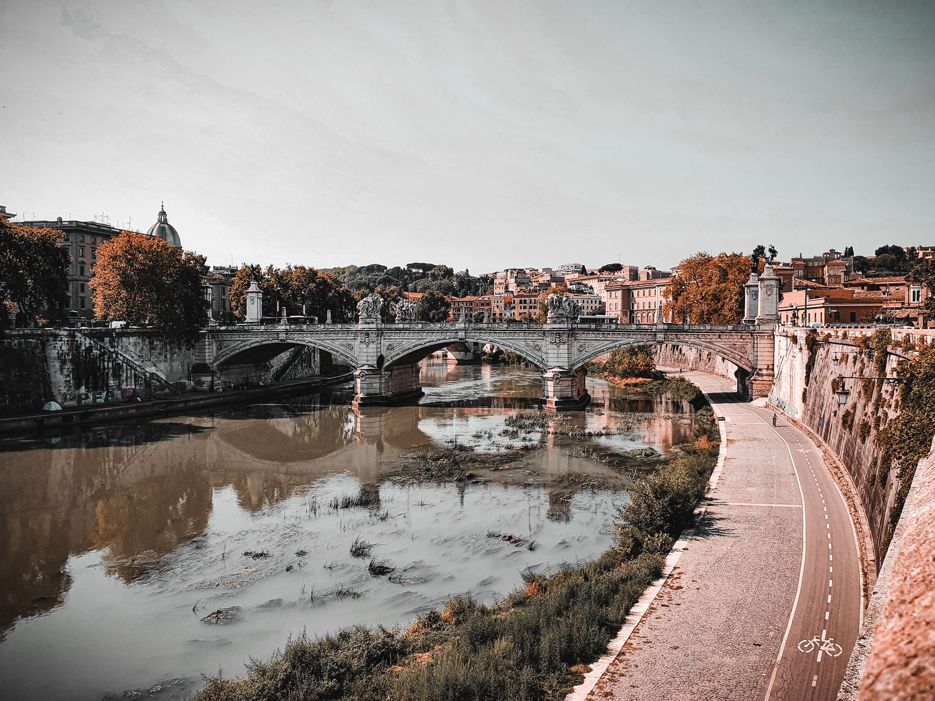 Ancient arched bridge with carved columns over Tiber river flowing along embankment in old city Rome