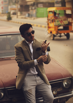 A man in a suit smoking, leaning on a vintage car in a street in Sialkot, Pakistan.