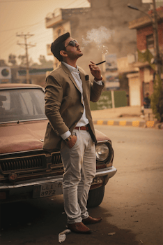 A man in a suit smoking a cigar while leaning on a vintage car in an urban setting.