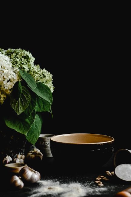 A rustic kitchen setup featuring a bowl, garlic, and hydrangea flowers against a black background.