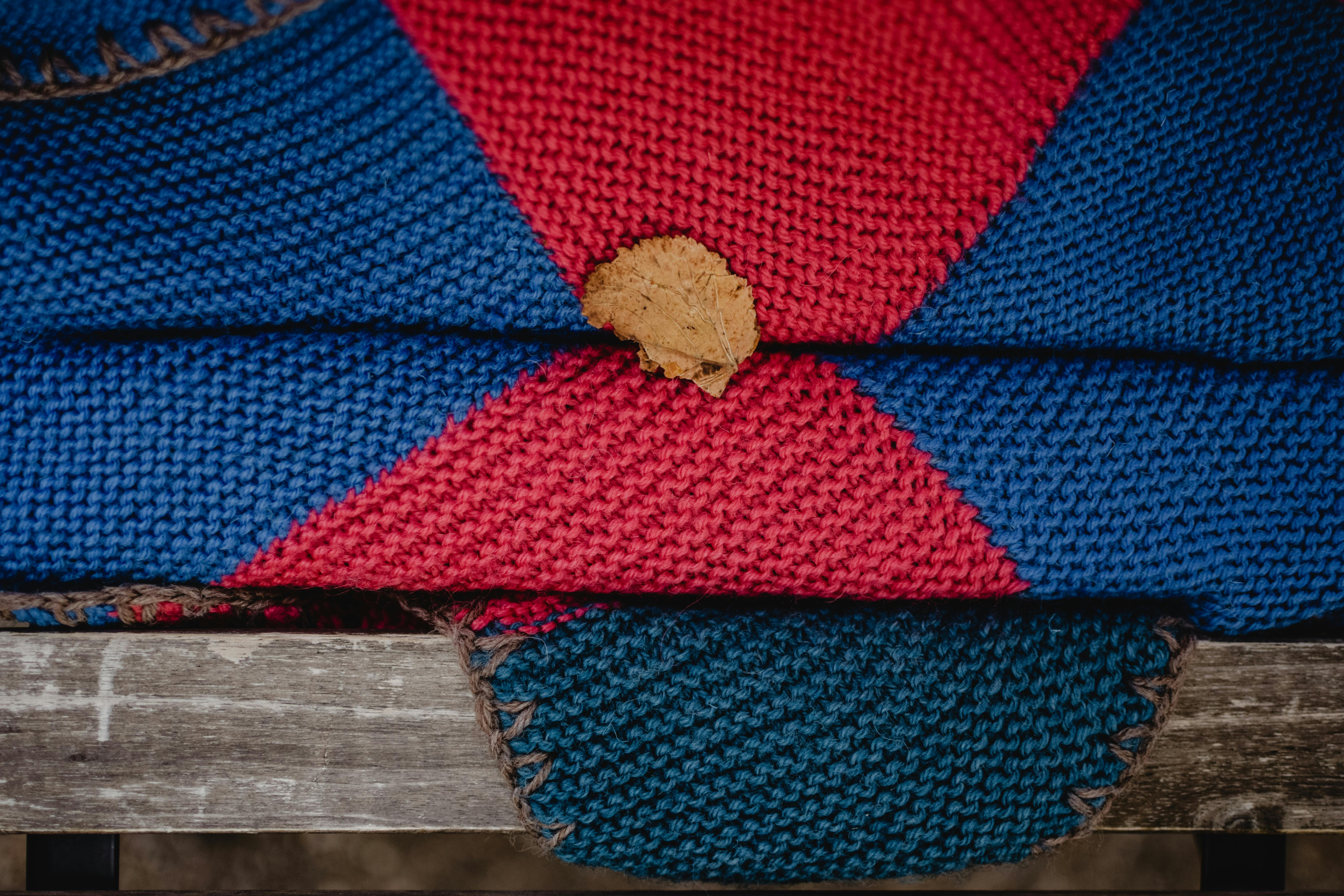 Handmade knitted blanket in vibrant red and blue featuring a fallen leaf on a wooden bench.