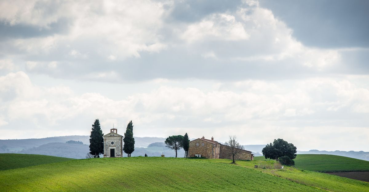 Charming Tuscan landscape with a chapel, farmhouse, and rolling green hills under a cloudy sky.