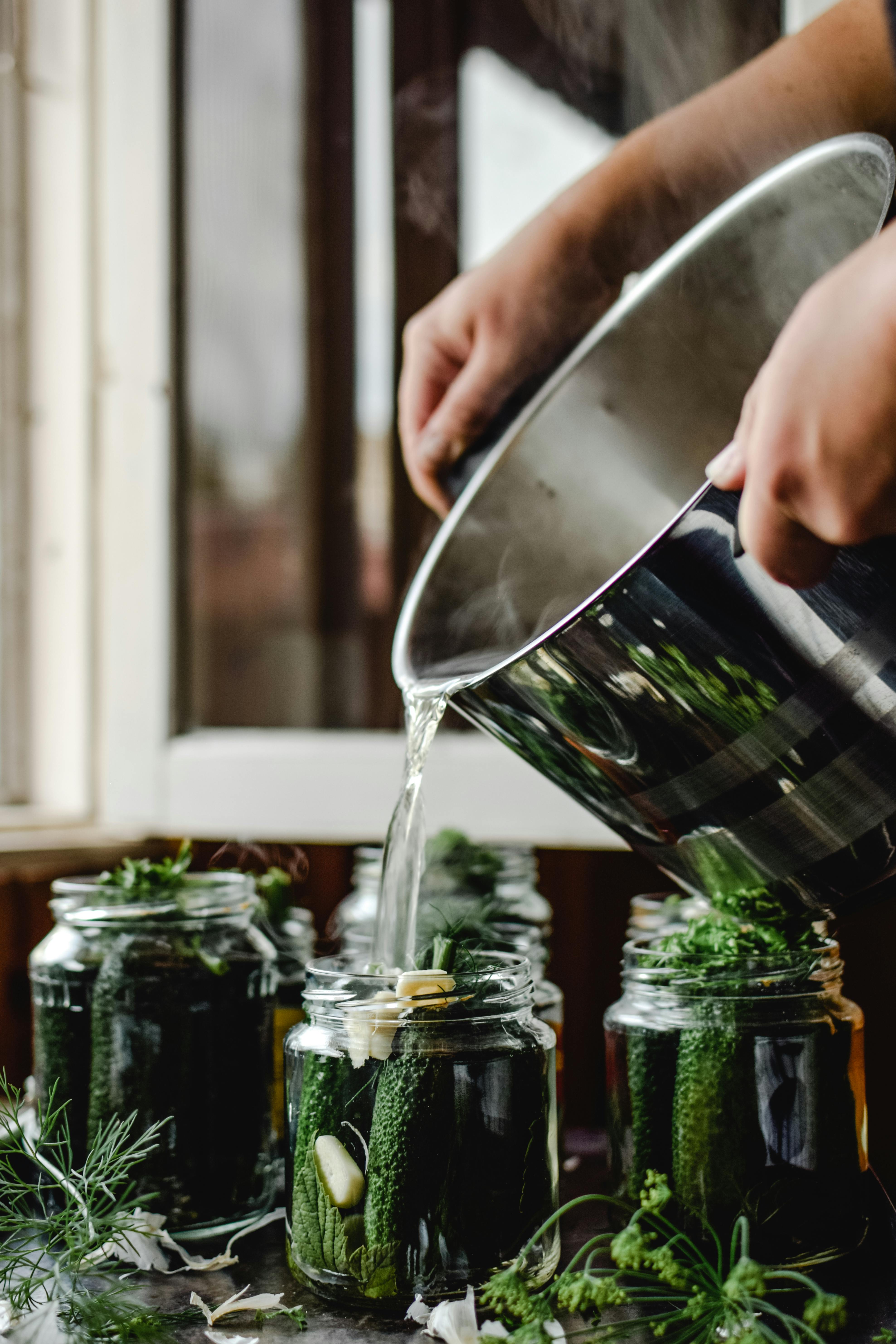 person pouring water on pickled cucumber jars