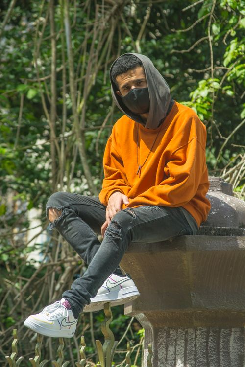 Free Young man wearing an orange hoodie and black mask sitting outdoors in a park on a sunny day. Stock Photo