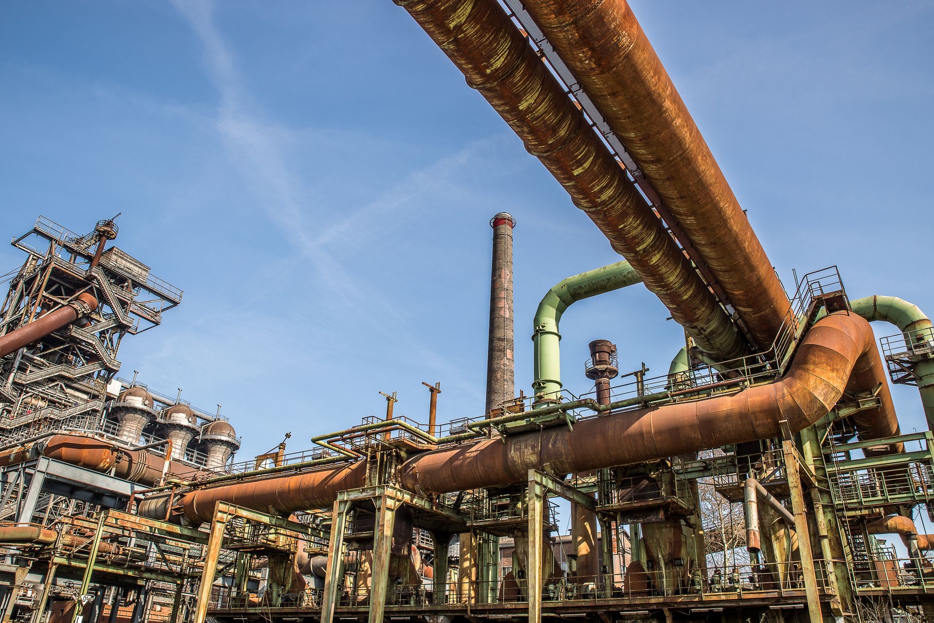 A detailed view of a rusty industrial facility with large pipelines and smokestacks under a clear blue sky.