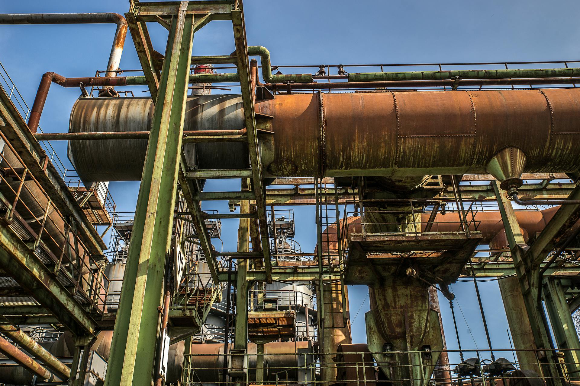 A low angle view of a rusty old industrial structure with pipes and beams against a clear blue sky.