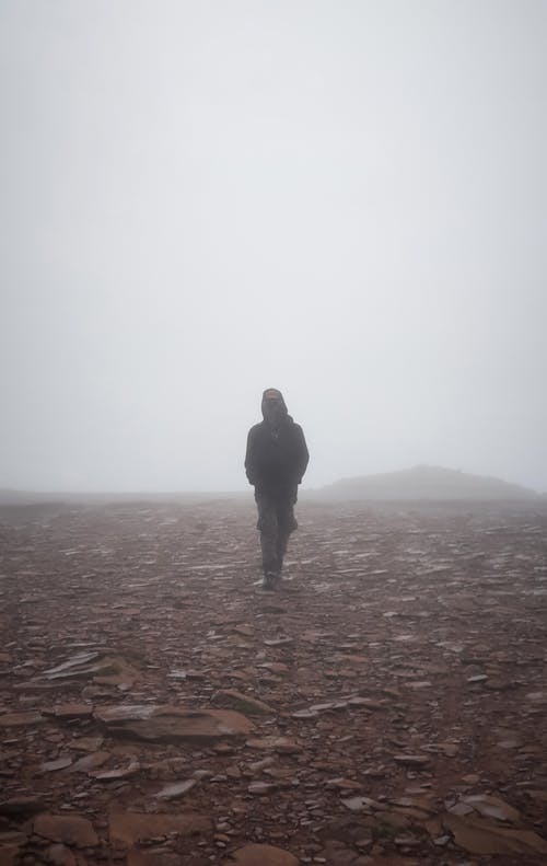 Free Full body of faceless male in hood holding hands in pockets and walking on dry stony ground in foggy weather Stock Photo