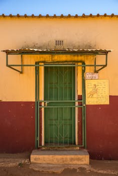 A rural school library entrance featuring a green metal door with protective bars under a corrugated iron roof.