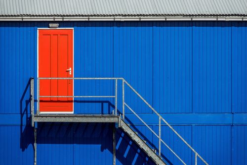 Free A striking contrast of a red door on a blue corrugated building facade with metal staircase. Stock Photo