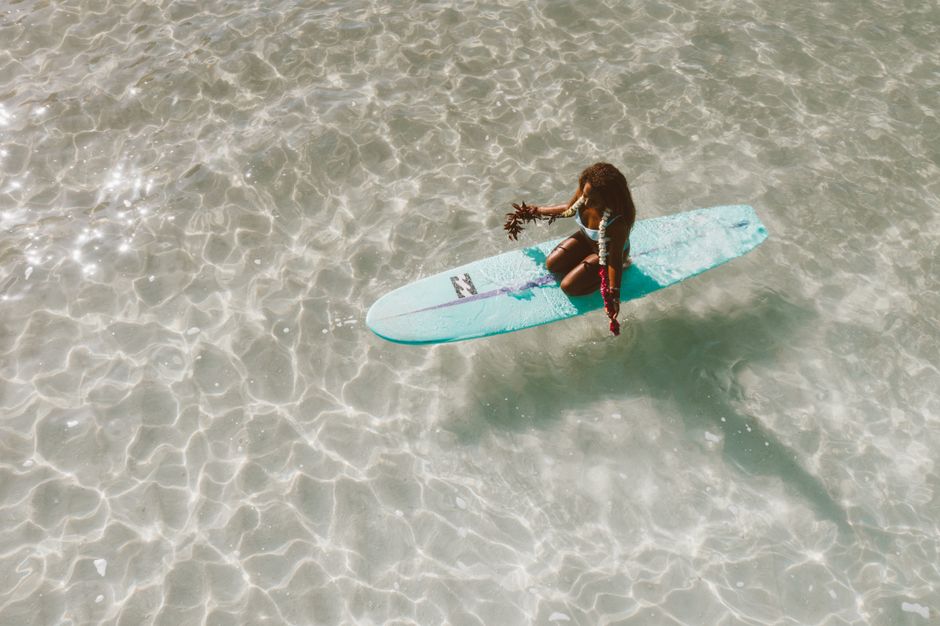 Woman on a surfboard holding seaweed in clear ocean waters, aerial perspective.
