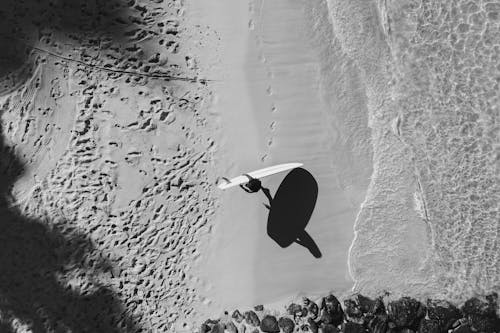 Monochrome Photograph of a Person Carrying a Surfboard at the Seashore