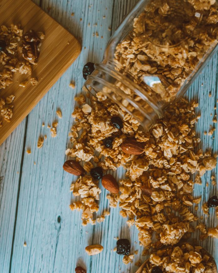 Granola clusters with almonds and dried fruit spilling from a glass jar onto a painted wood table, photo by Jess Loiterton on Pexels