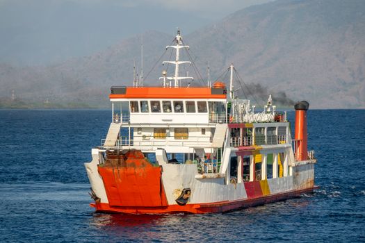 A colorful ferry navigates the open sea under clear daylight, with scenic mountains in the background.