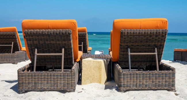 Wicker lounge chairs with orange cushions facing the sea on a sunny beach.