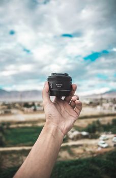 Close-up of a hand holding a Canon lens against a scenic sky and blurred landscape.