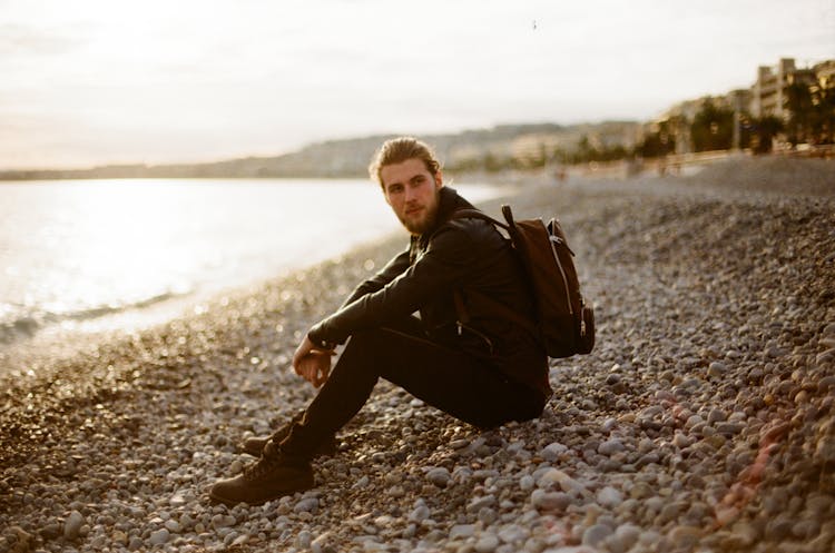 A Man In Black Jacket Sitting On The Rocky Shore Of A Beach