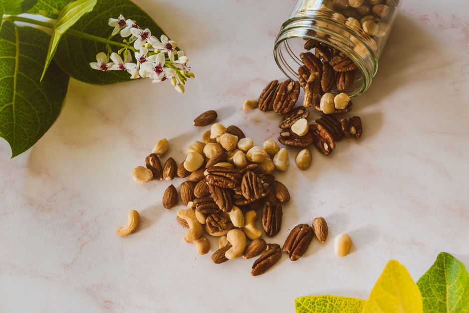 Hạt sang - bí mật chữa dạ dày của người Mông 3 Assorted nuts spilling from a clear jar onto a marble surface with green leaves and white flowers.