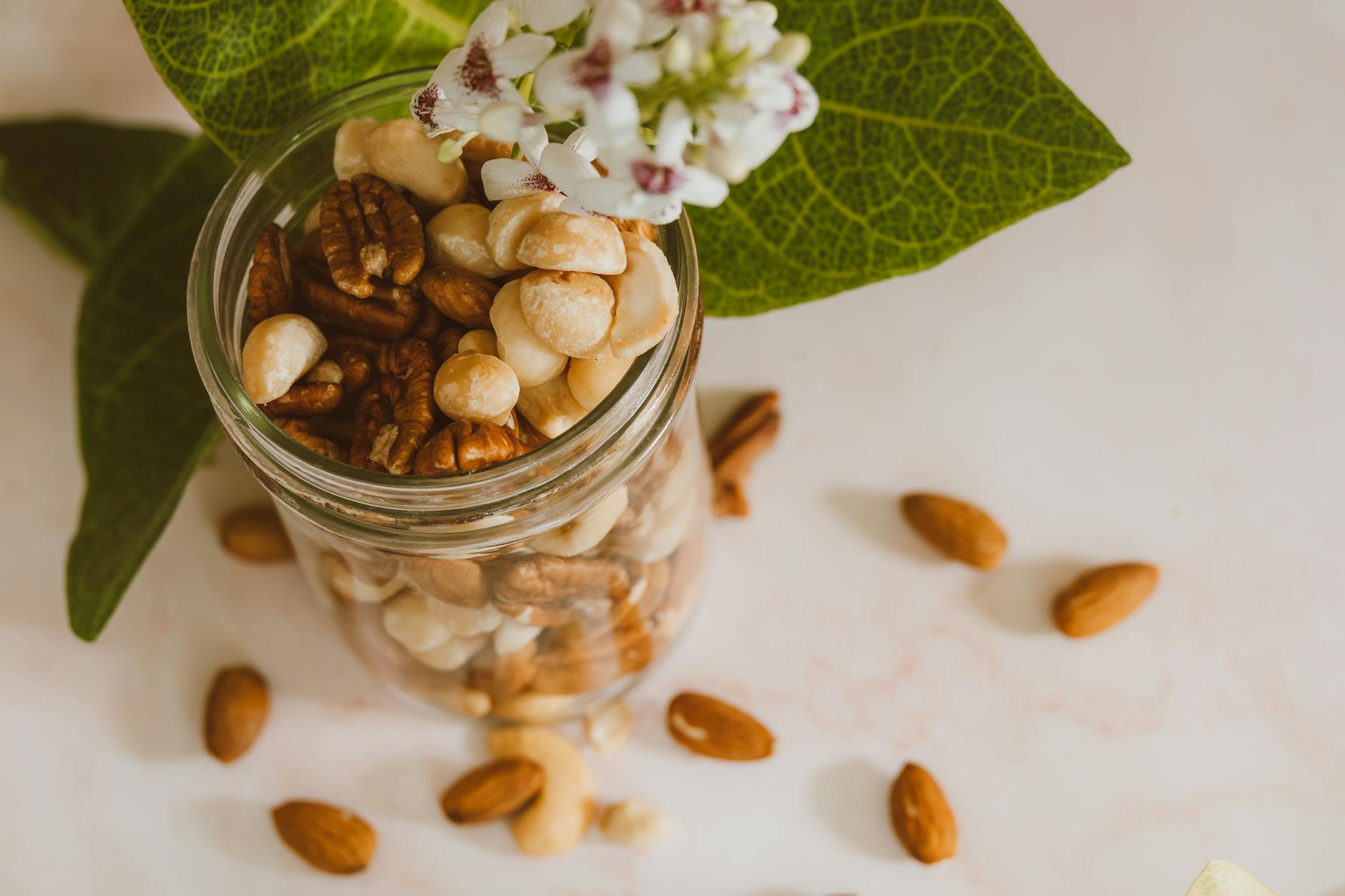 Assorted organic nuts in a clear glass jar with vibrant green leaves and white flowers for a healthy lifestyle.
