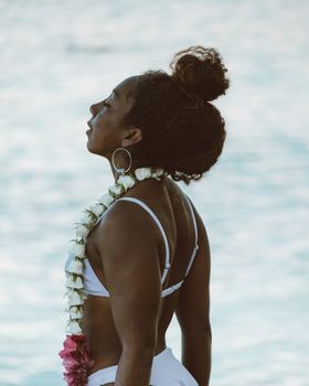 A woman in a white bikini poses gracefully by the ocean, adorned with a floral lei.