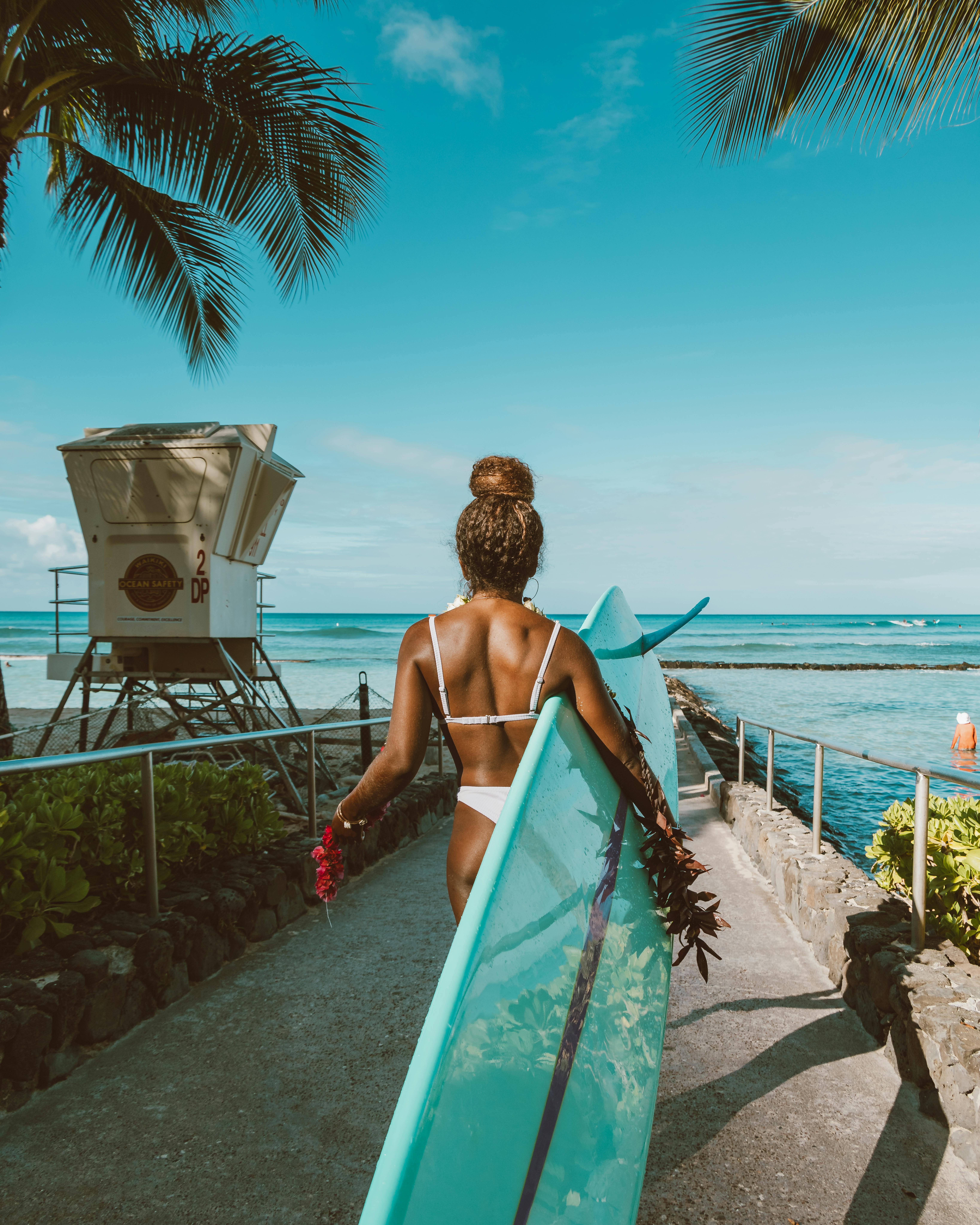 一名女子背着冲浪板，沿着阳光明媚、长满棕榈树的热带海滩小路走过