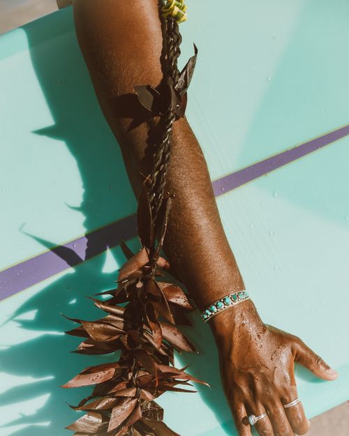 Free Close-up of a sunlit arm holding a surfboard, adorned with leaves and bracelets. Stock Photo