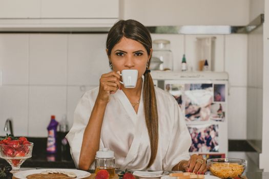 A woman in a white robe enjoys coffee with breakfast in a cozy kitchen setting.