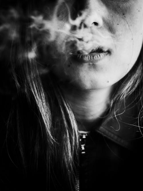 Black and white crop thoughtful sad female smoker exhaling smoke while depressing and spending time alone