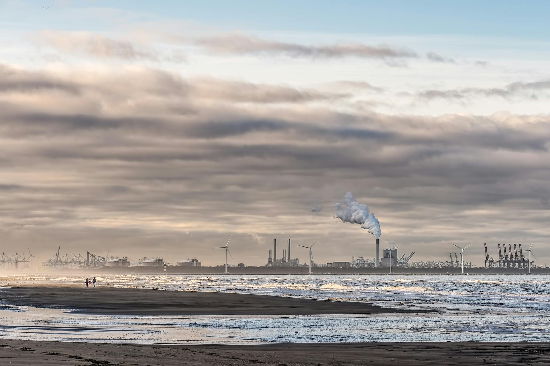 A coastal view of an industrial plant with smokestacks and wind turbines by the beach.