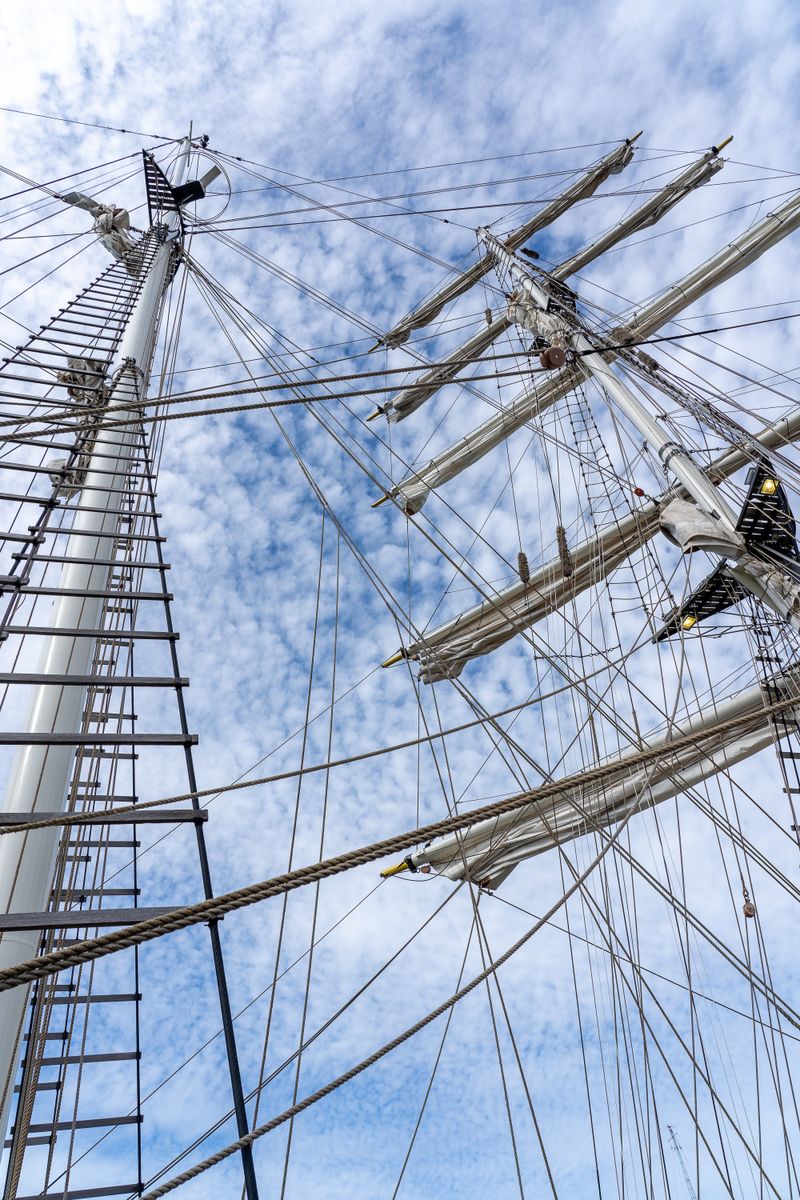 Looking up at a tall ship's masts and rigging against a bright blue sky with scattered clouds