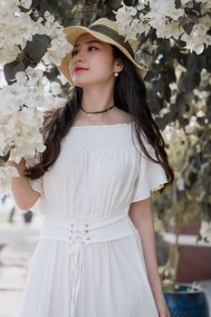 一名女子身穿时尚白色连衣裙、头戴帽子，在户外的九重葛花旁优雅地摆着姿势
