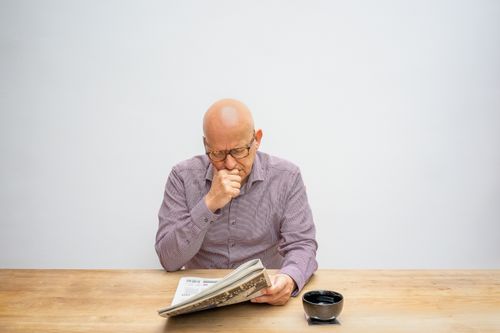 Free Bald senior man wearing eyeglasses reads a newspaper thoughtfully at a wooden table with a cup. Stock Photo