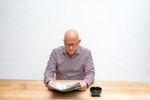 Free Bald senior man with eyeglasses reading a newspaper at a wooden table. Stock Photo