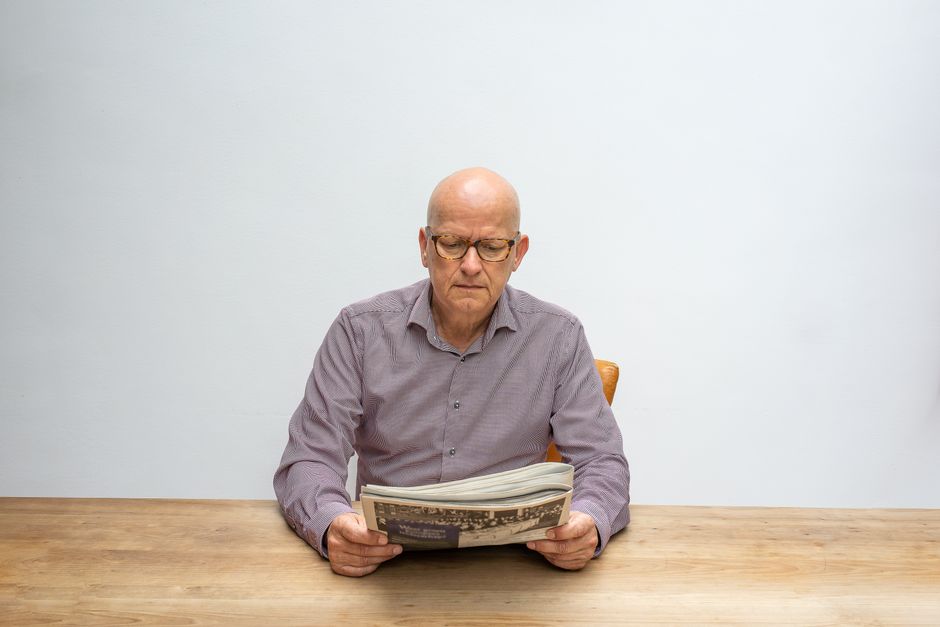 Senior man with eyeglasses reading a newspaper at a wooden table against a white background.