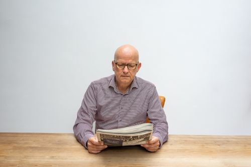 Free Senior man with eyeglasses reading a newspaper at a wooden table against a white background. Stock Photo