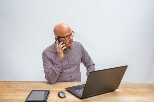 Free Senior man wearing eyeglasses using cellphone while working on laptop, illustrating modern business communication. Stock Photo