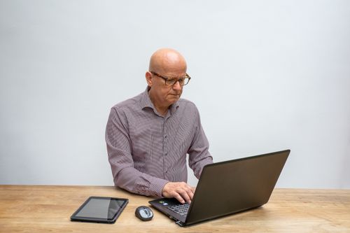 Free Elderly man working on laptop with tablet and mouse on table in a light room. Stock Photo